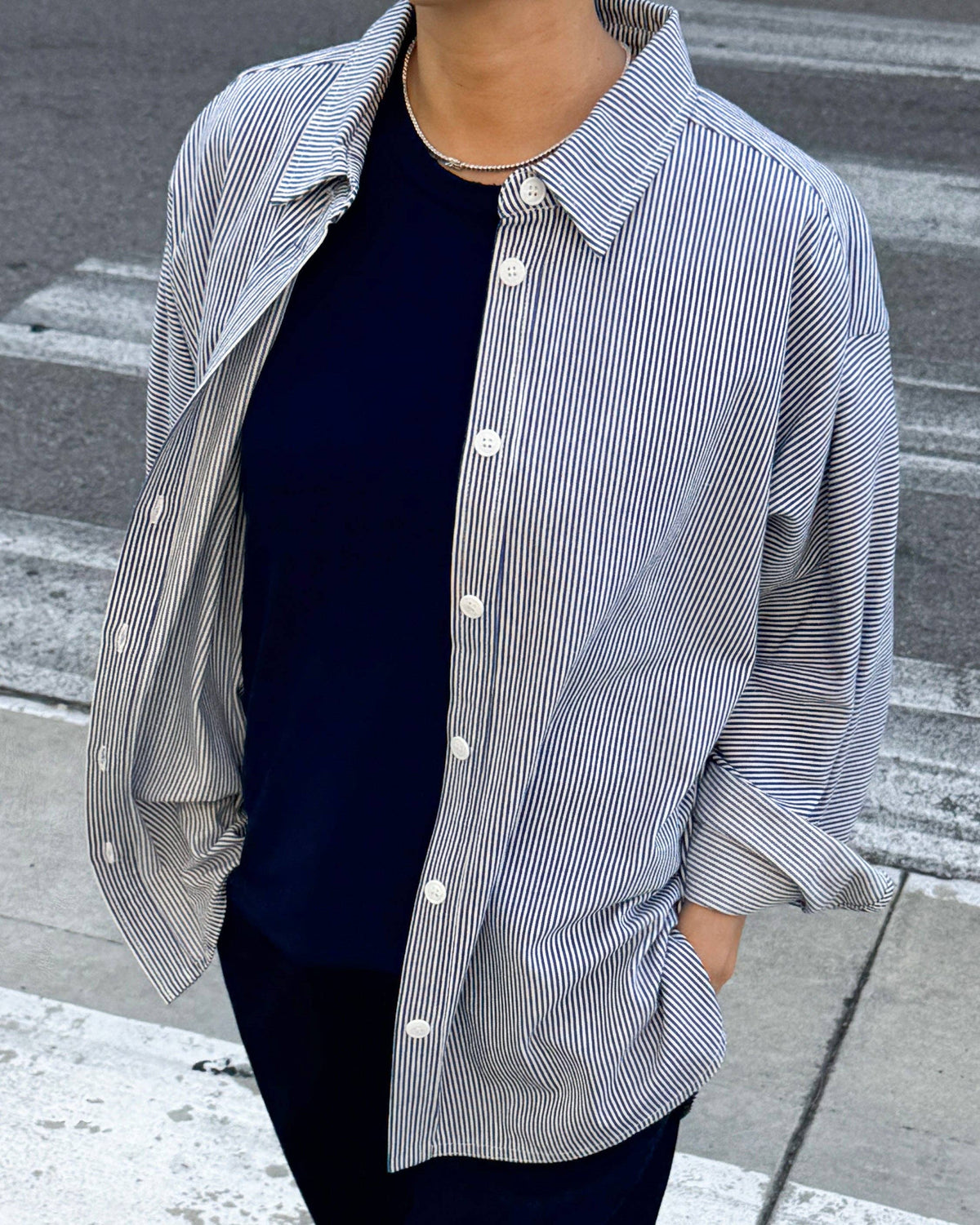A person wearing an oversized long-sleeve button-up shirt with fine vertical blue and white stripes, layered over a dark navy crewneck top and dark pants, standing on a city crosswalk.