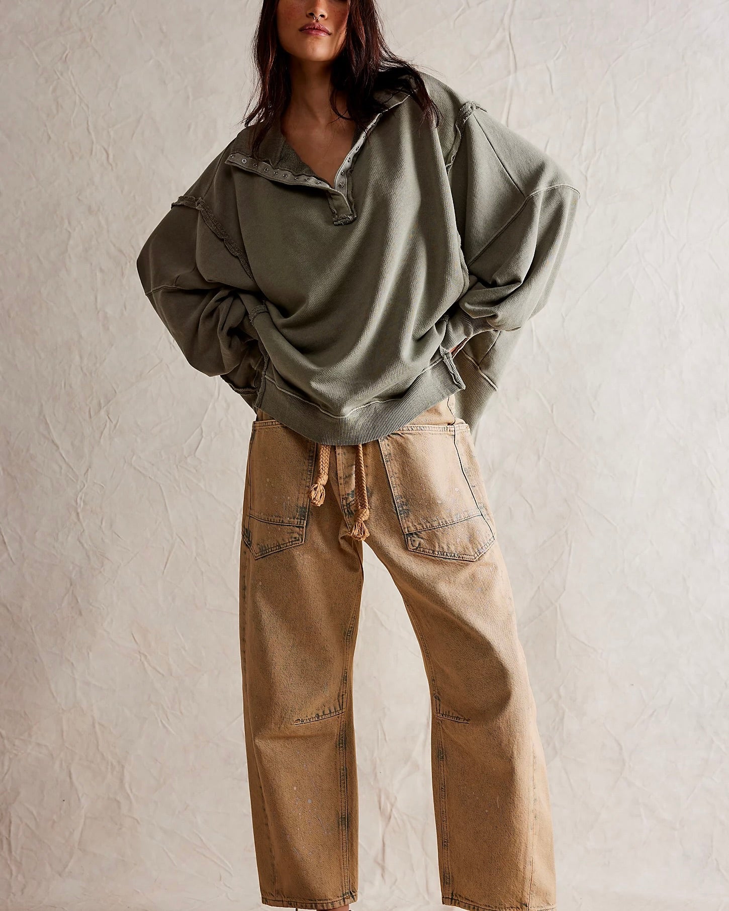 Model wearing Free People Camden Henley sweatshirt in Laurel Oak with tan pants standing in front of a white wall.