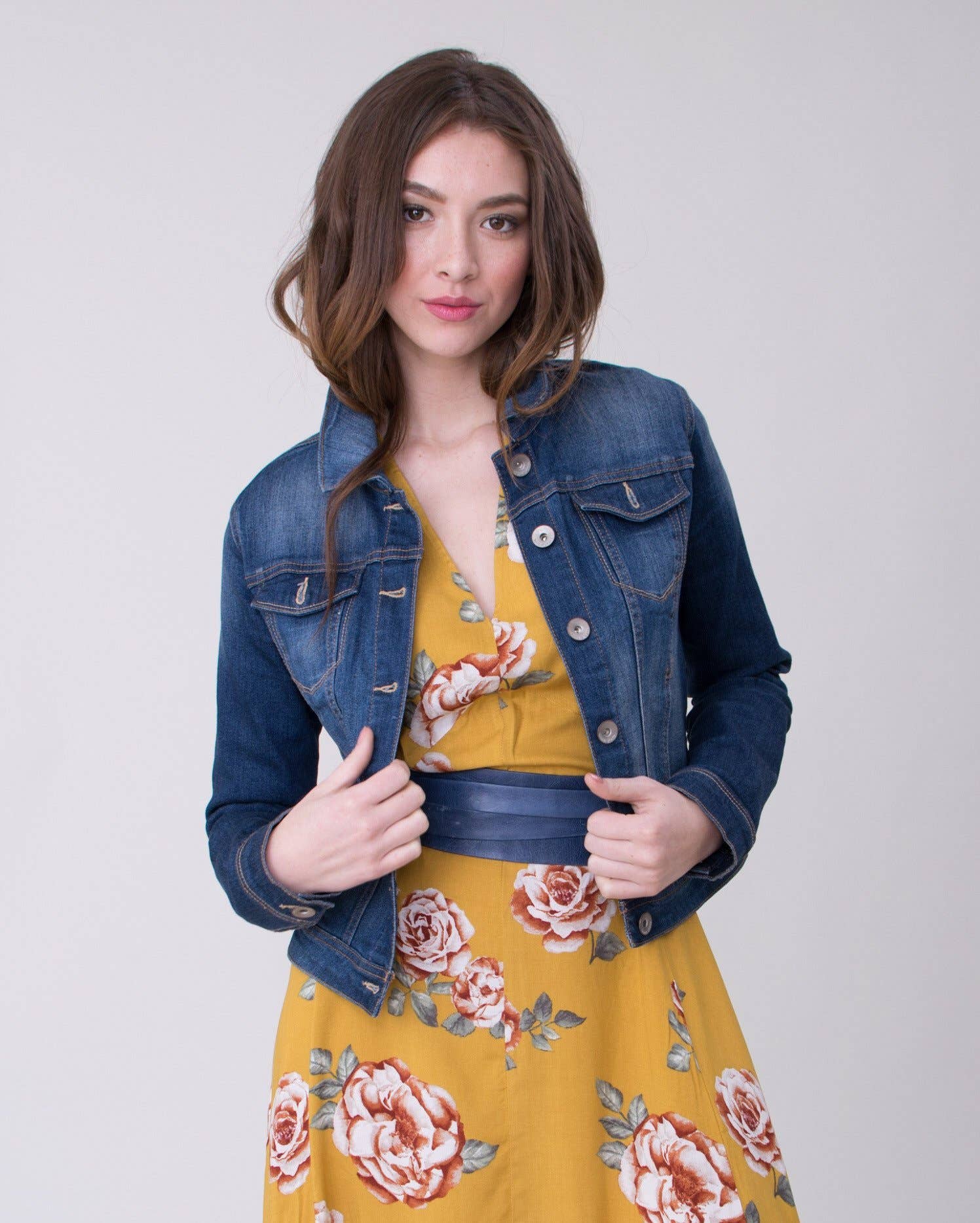 Woman wearing a denim jacket over a yellow floral dress with a navy wrap belt on a plain background