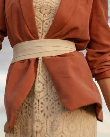 Person wearing a rust-colored jacket over a beige patterned dress with a belt.
