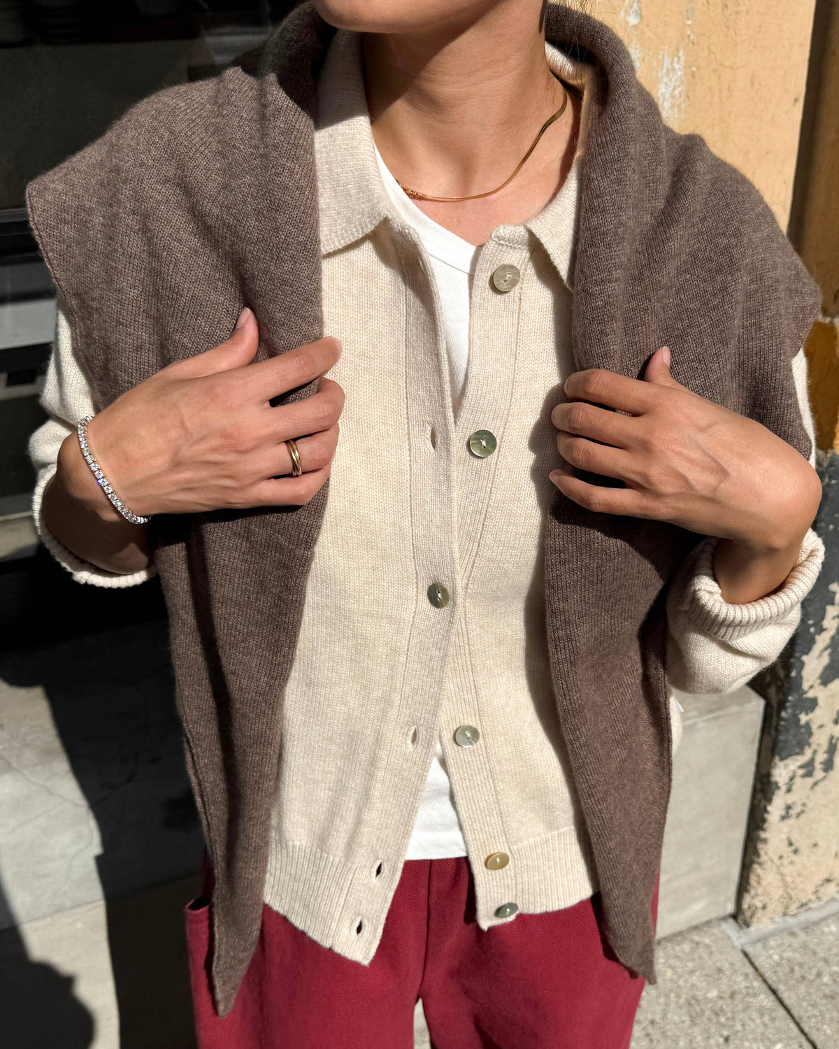 A person outdoors wearing a brown knit sleeveless cardigan layered over a cream button-front cardigan, a white top, and burgundy pants.