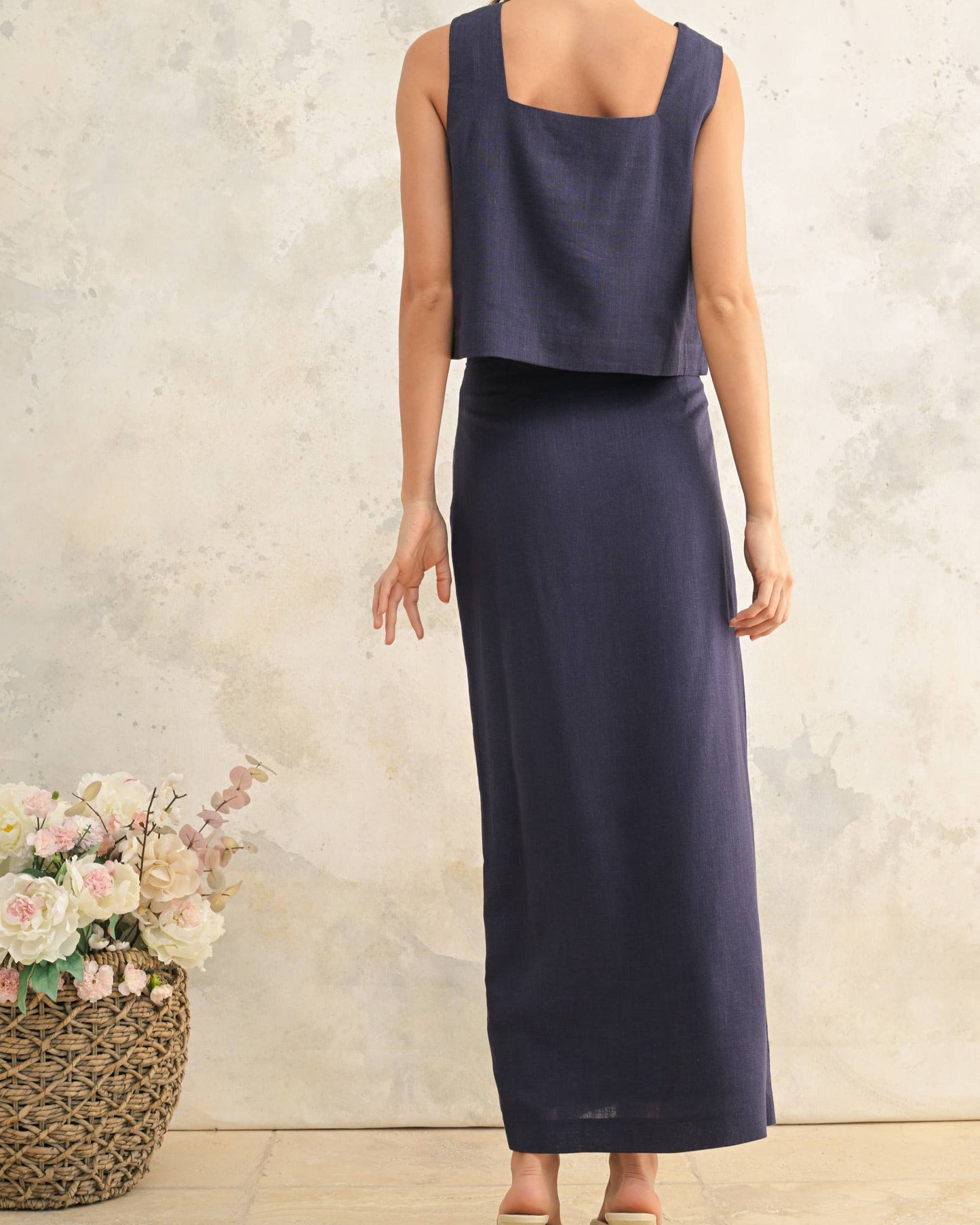 Woman wearing a navy blue top and skirt standing against a textured wall with flowers in the foreground.