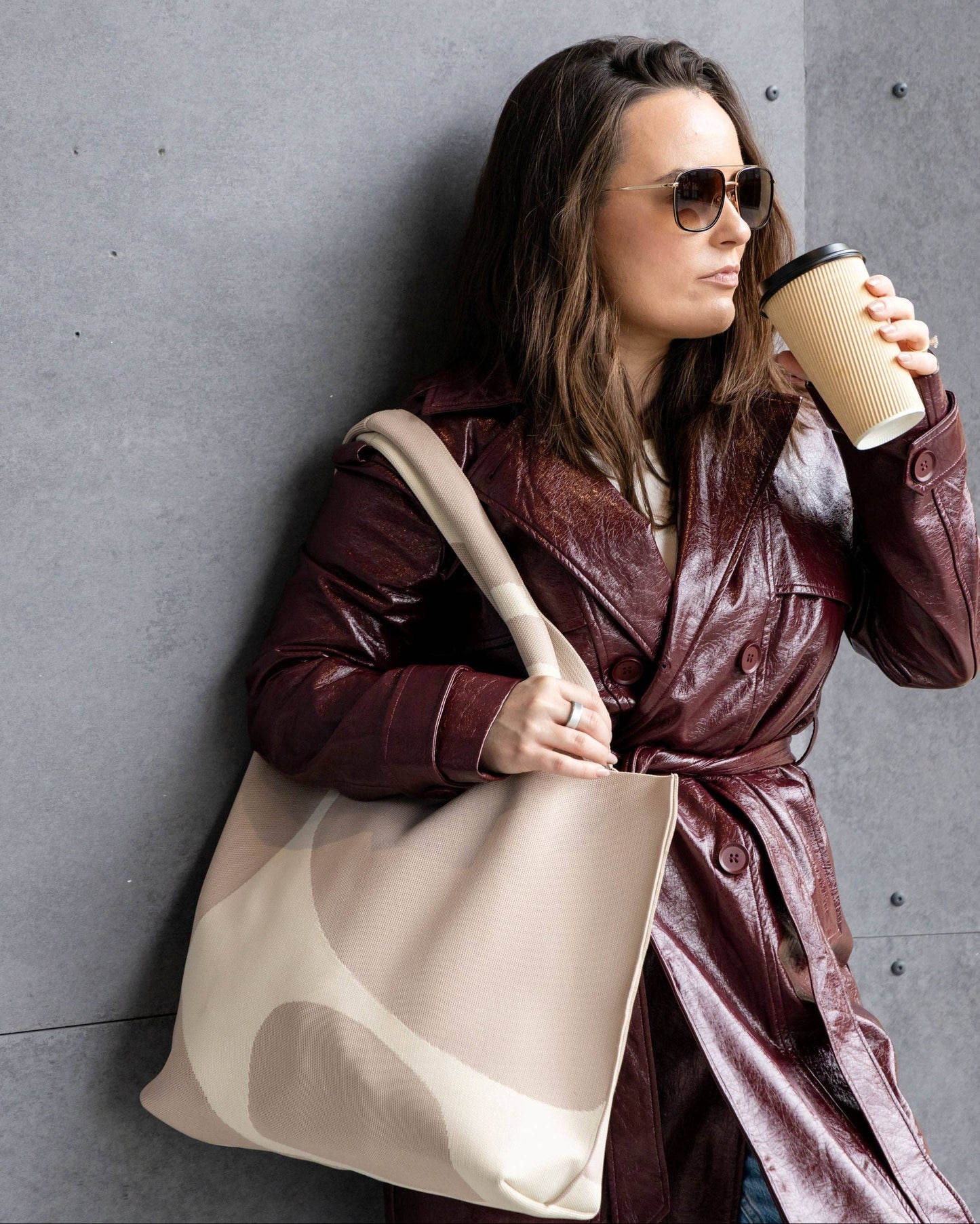 Woman in a burgundy coat holding a beige tote bag and a coffee cup against a gray wall.
