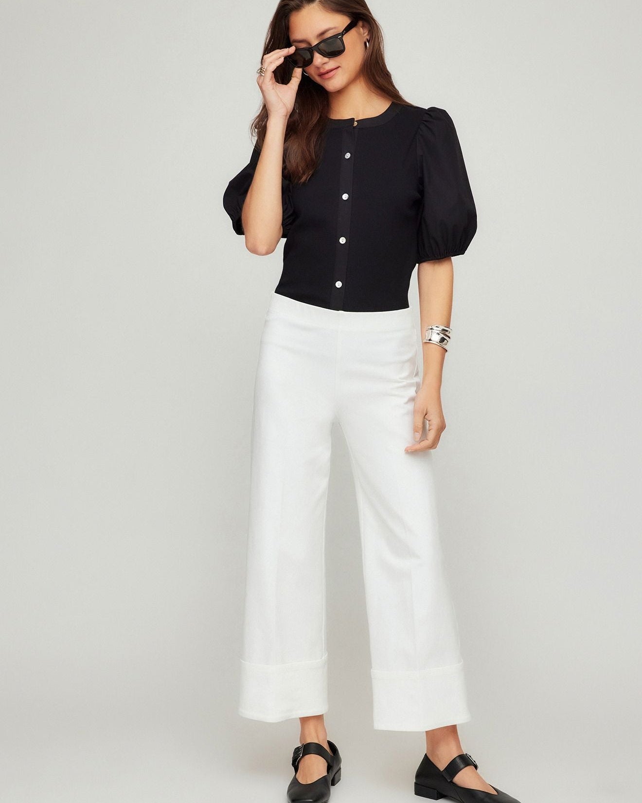 Woman wearing a black top and white pants with sunglasses on a plain background