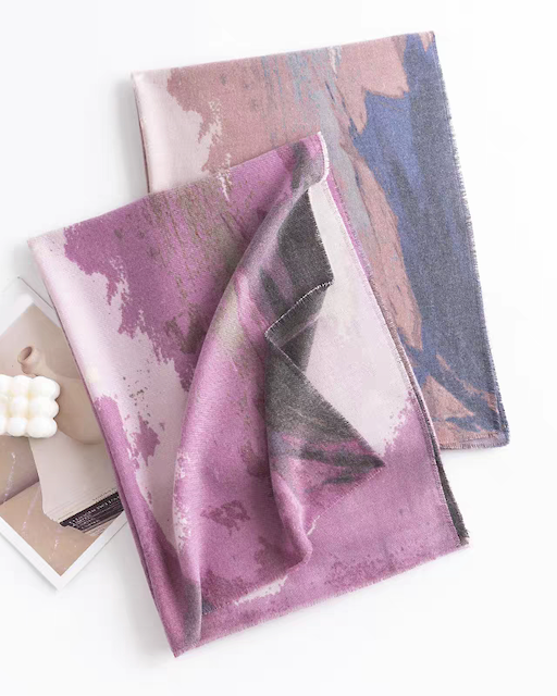 Two abstract patterned scarves in shades of purple, pink, and blue-gray are arranged on a white background, with one folded over the other. A small printed card and a white decorative object are partially visible underneath.
