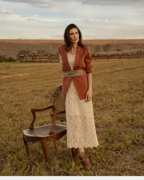 Woman in a rust-colored jacket and white lace dress standing in a field with a wooden chair.