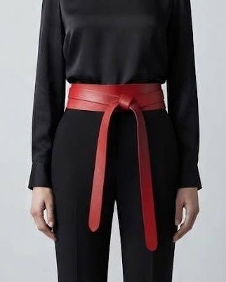 A model wearing a black long-sleeve top and black trousers, styled with a wide, bright red leather belt tied at the waist, set against a plain light gray studio background.