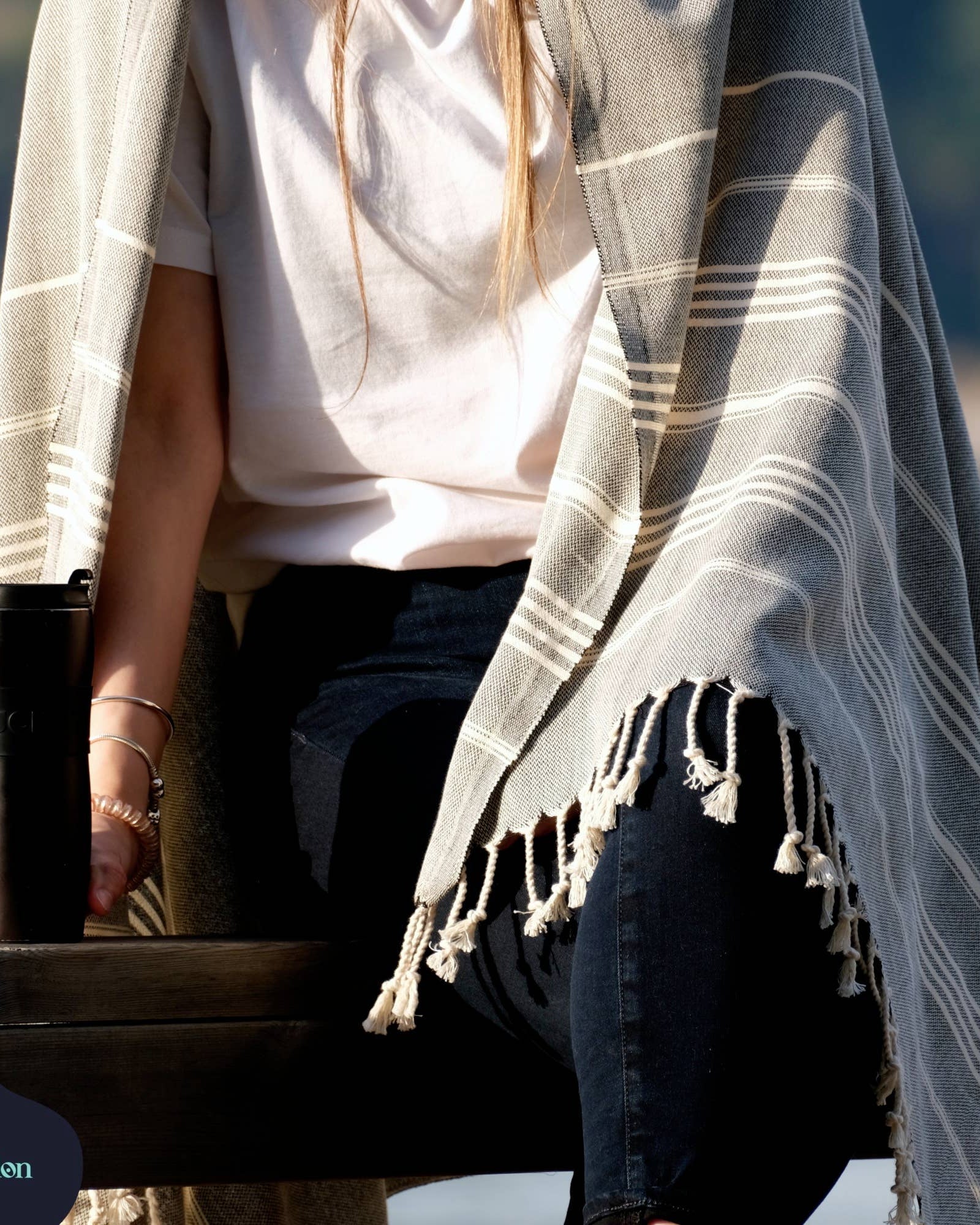 Person holding a tumbler with a plaid shawl draped over their shoulders by a body of water.