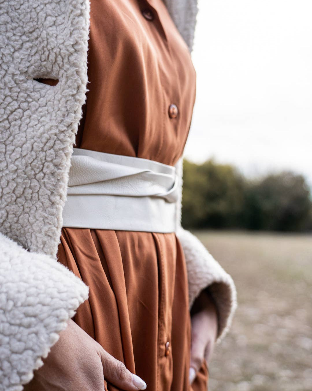 Person wearing a brown coat with a white shirt and beige cardigan in an outdoor setting