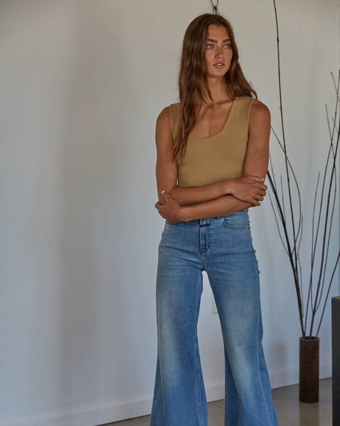 Woman wearing a beige tank top and blue jeans standing indoors with a minimal background