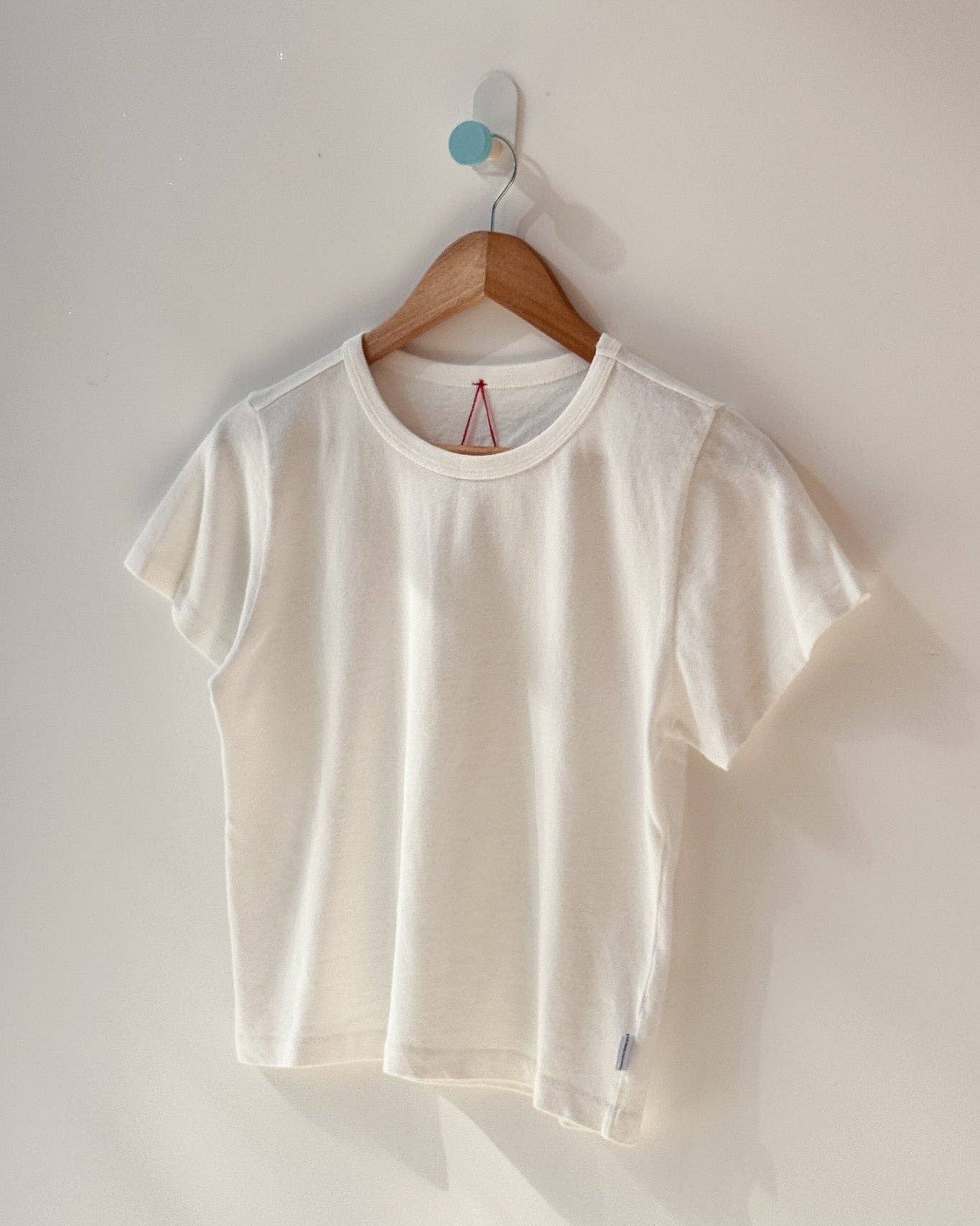 A white short-sleeve crewneck t-shirt hanging on a wooden hanger from a wall hook against a plain off-white wall.