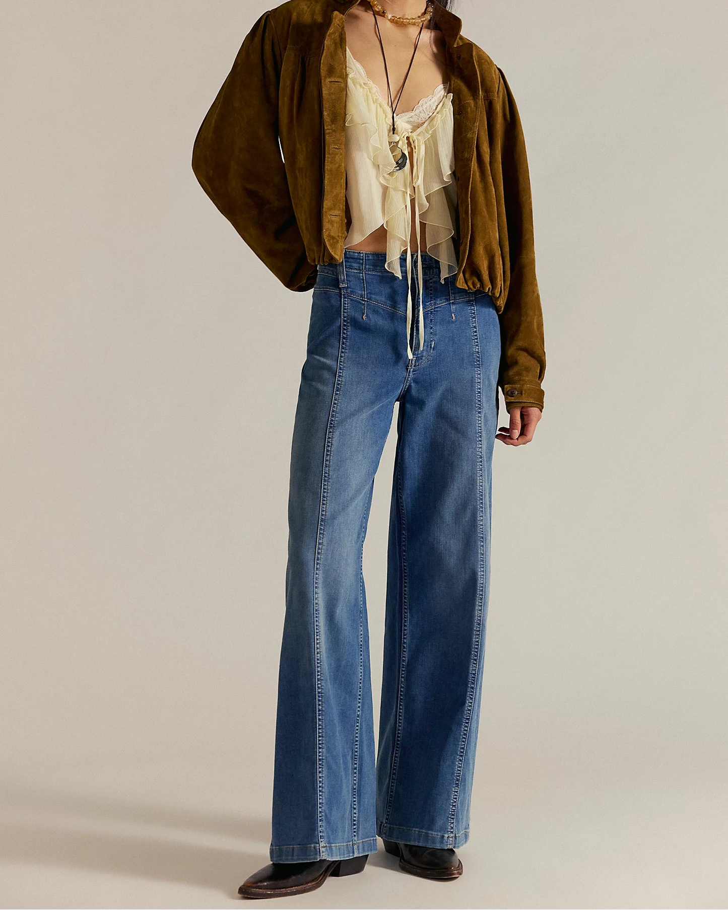 Woman wearing a brown jacket, beige top, and blue jeans on a plain background