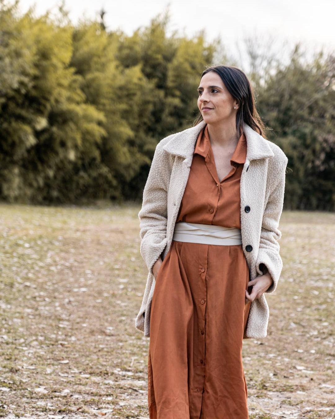 Woman in a rust-colored dress and beige coat standing in a field with trees in the background