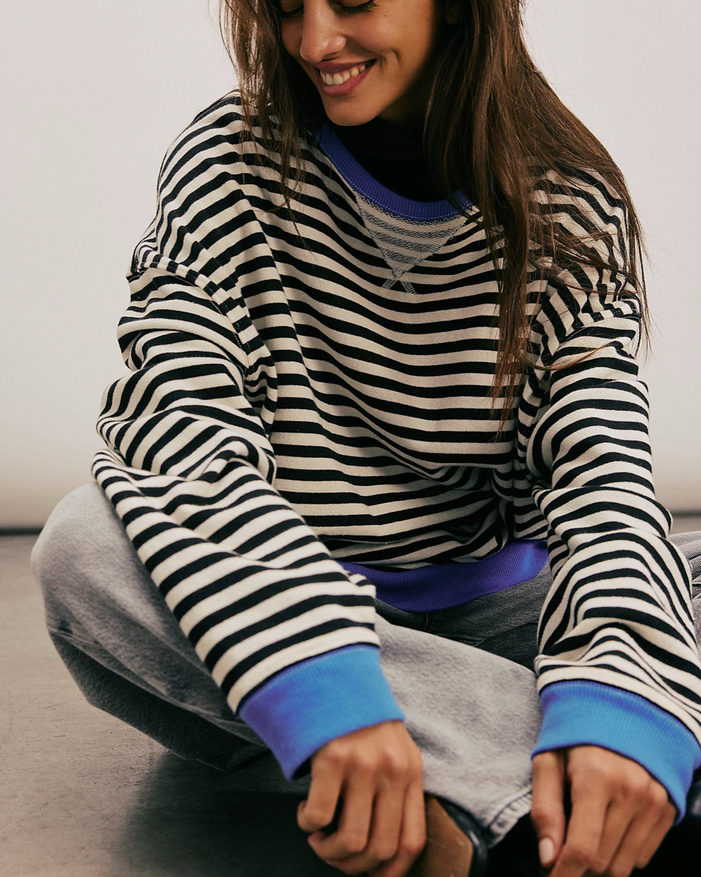 Front view of model sitting down, wearing the Free People Classic Striped Oversized Crewneck in front of a white wall. 