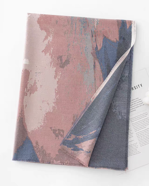 A folded abstract-patterned scarf in dusty pink, slate blue, and charcoal gray, styled on a white surface next to a magazine page and a small white pom-pom accessory.