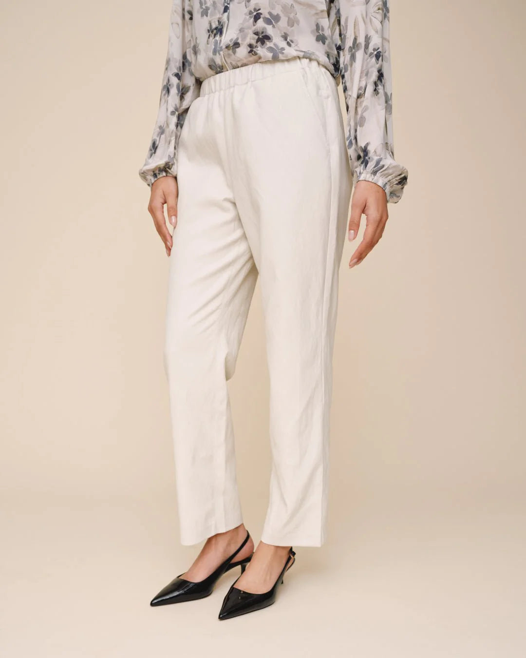 Person wearing a floral blouse, white pants, and black shoes on a beige background