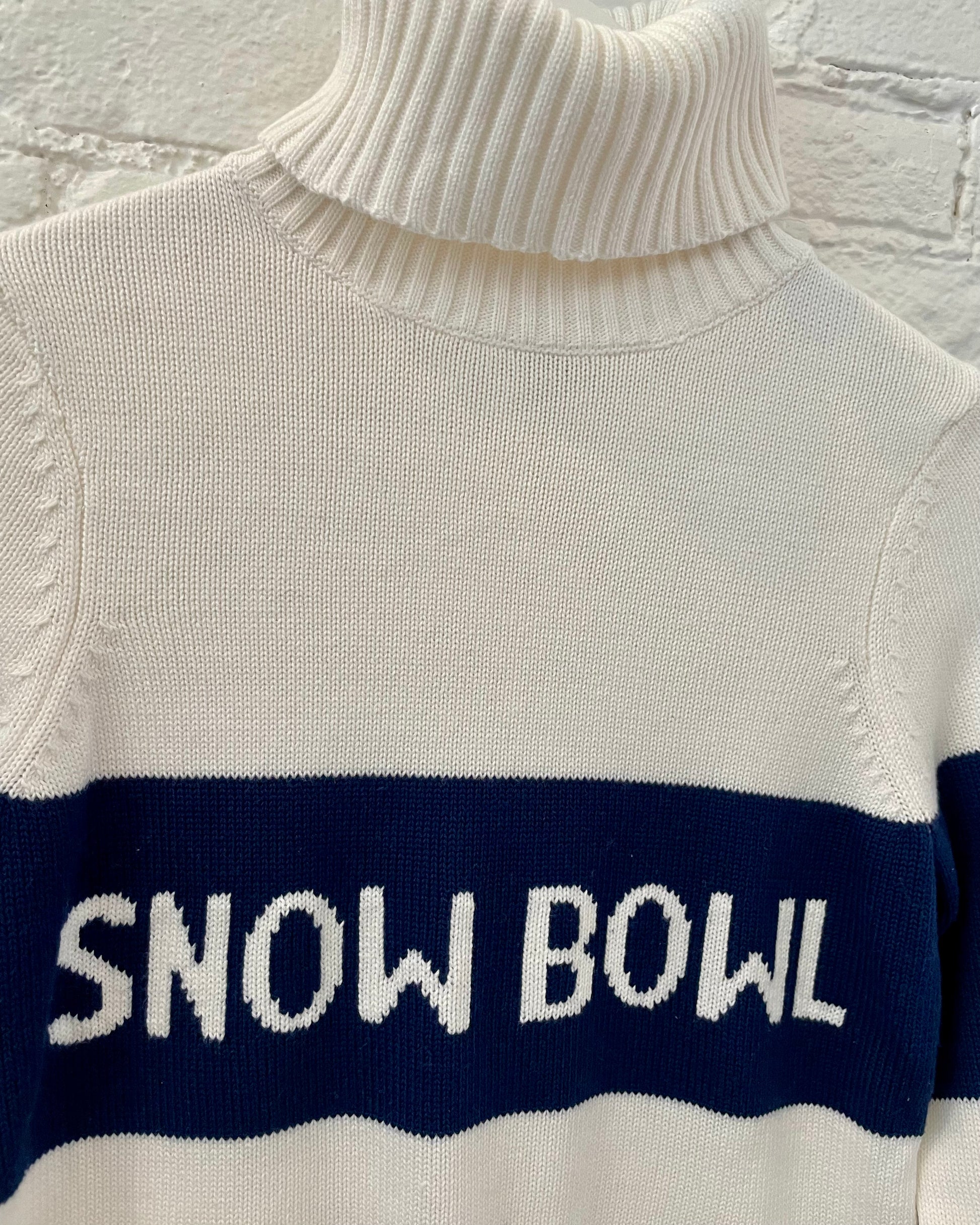 close up of ivory turtleneck sweater with navy blue stripe that reads snow bowl in ivory lettering 