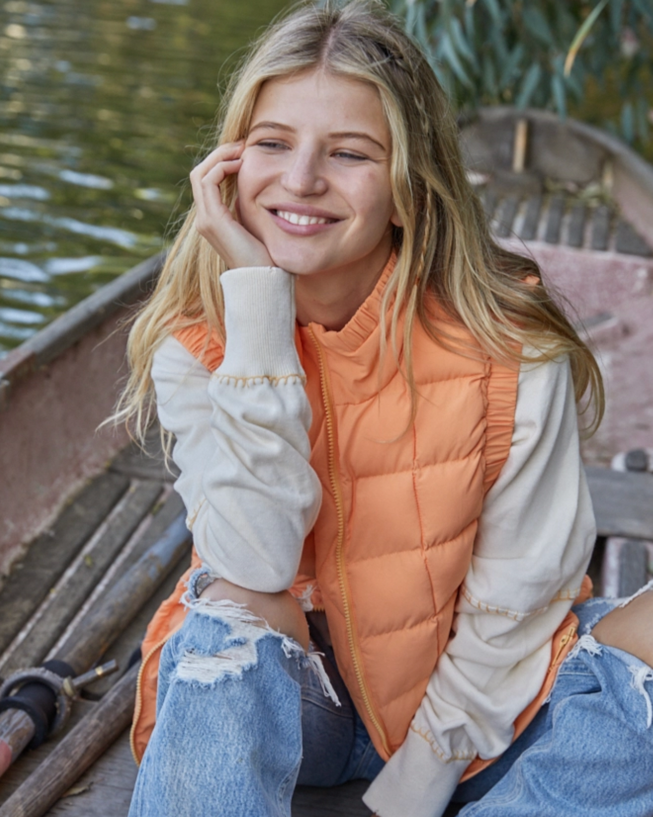Model wearing By Together Cool Orange Edeline Packable vest wearing jeans sitting by a lake 