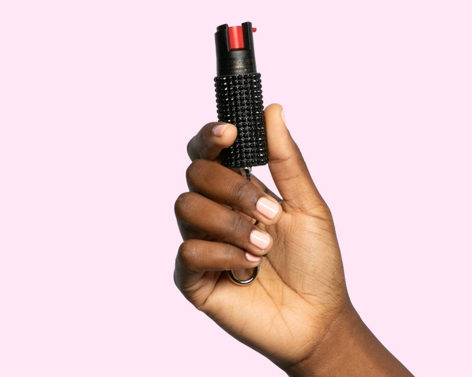 Image of a hand holding  Blingsting Pepper Spray in Black on a lavender background