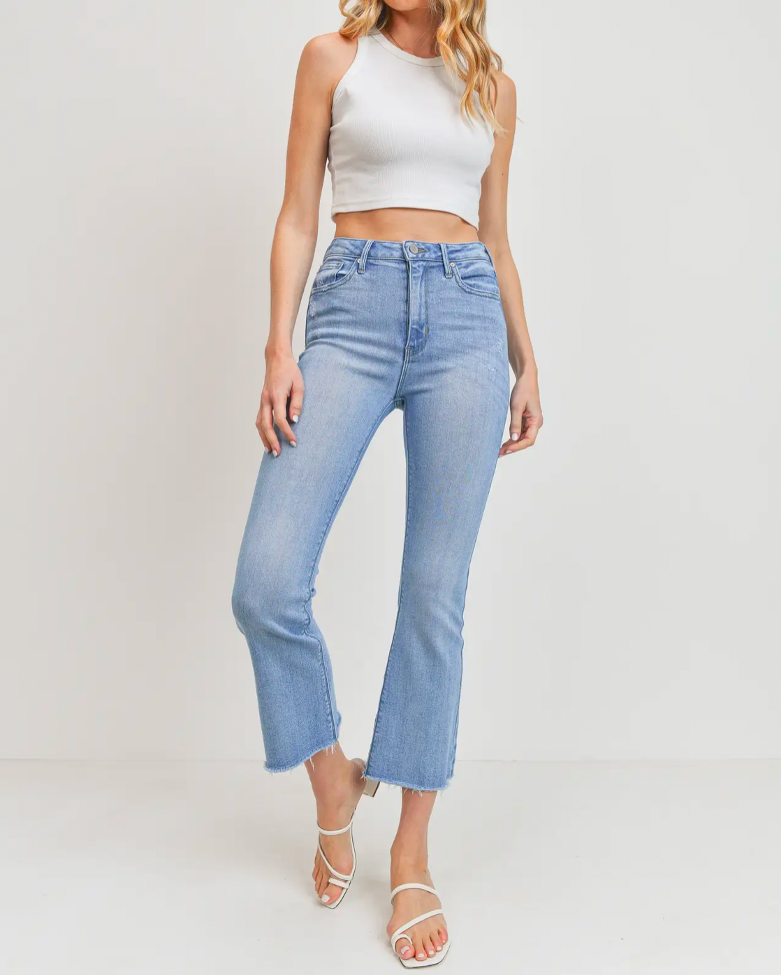 Model wearing Just black denim High Rise Tonal Crop Flare Light wash jeans wearing white crop tank top and white sandals on a white background