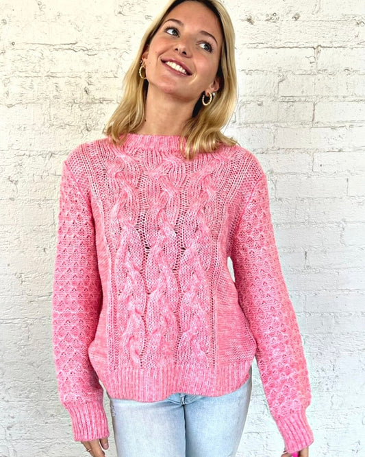 Model wearing Wooden Ships Mitch Cable Crew Pretty in Pink Sweater wearing jeans standing behind a white brick background