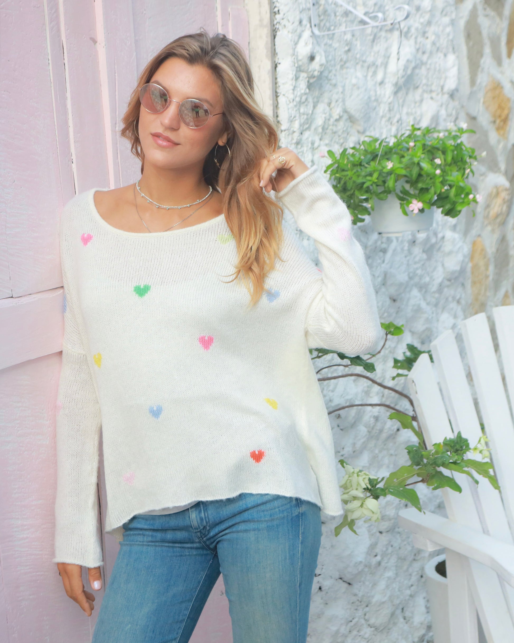 Model wearing Wooden Ships Rainbow Hearts Crew sweater wearing jeans and sunglasses standing outside against a pink door