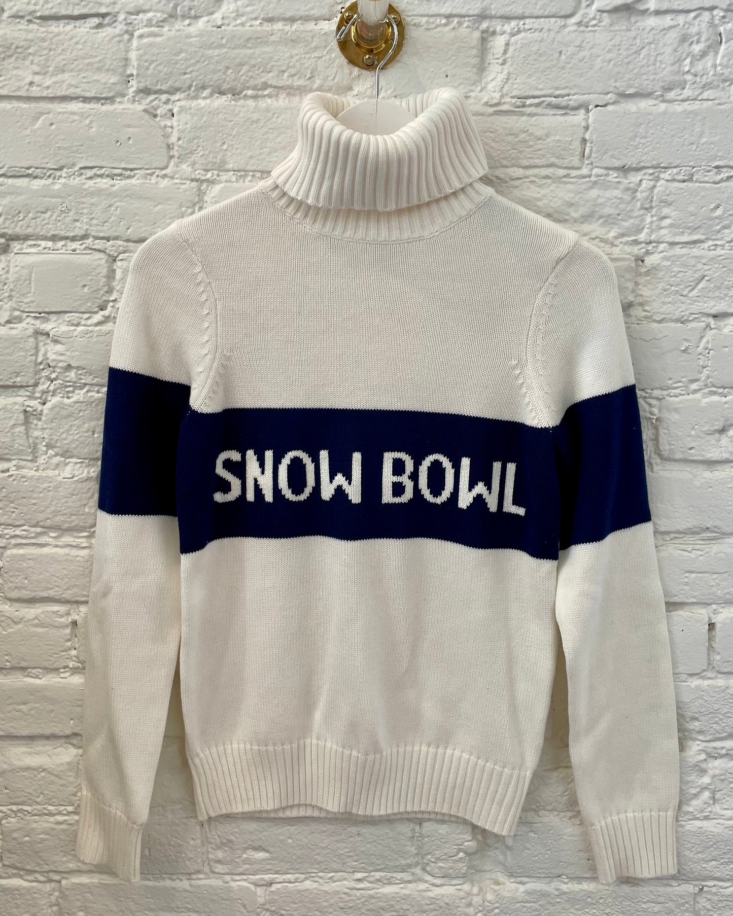Image of SNOW BOWL ivory turtleneck sweater with navy stripe that reads snow bowl in ivory lettering hanging on white brick wall 