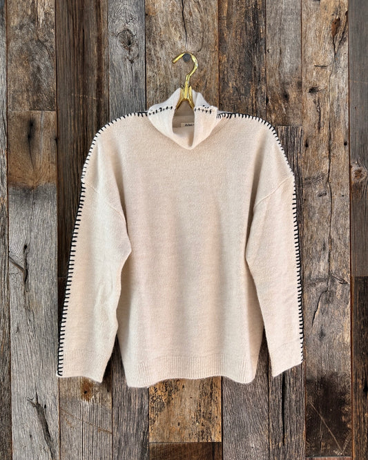 Image of Dylan Daisy Earth Sweater-oatmeal with black stitching hanging on a hanger on a wooden wall