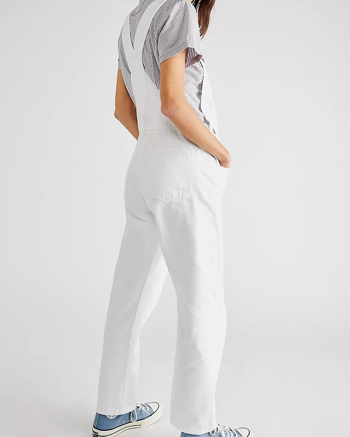 Model wearing Free People Ziggy Denim Overalls in white wearing blue shoes and a black and white stripe shirt on a white background
