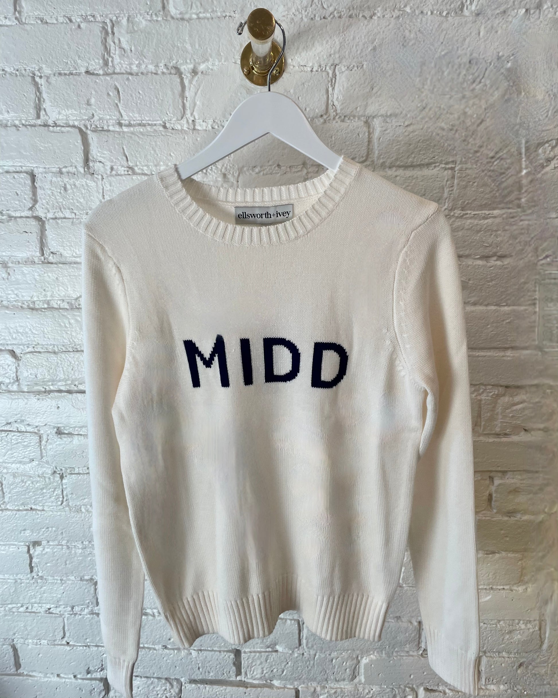 Middlebury College MIDD white sweater with blue letters from Ellsworth+Ivey on a white brick wall background