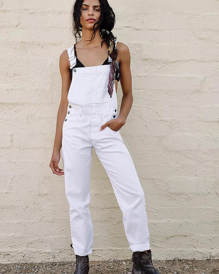 Model Wearing Free People Ziggy Denim overalls in Optic white wearing black boots and black tank on a cream brick background