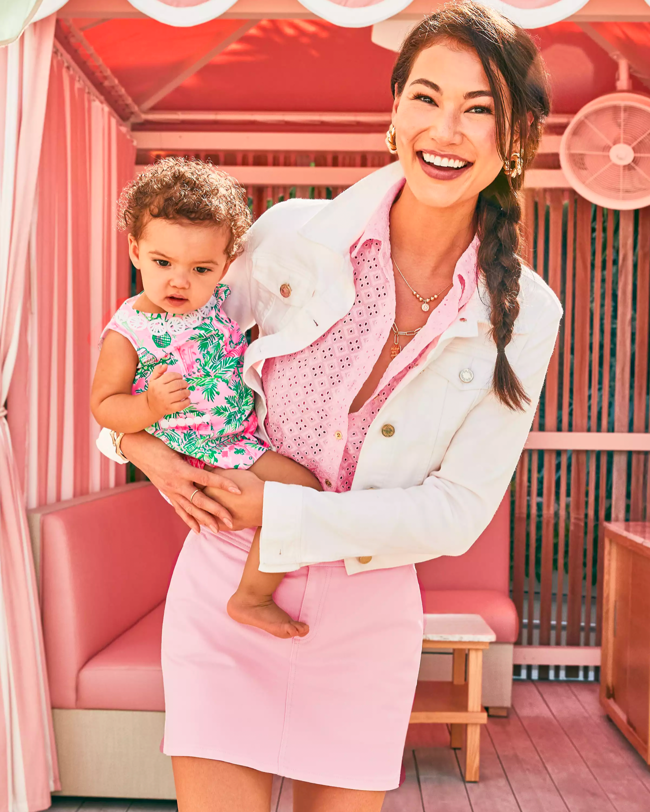 Model wearing Lilly Pulitzer Laylani Denim jacket in resort white holding a baby wearing pink skirt and pink top standing in front of a pink couch 