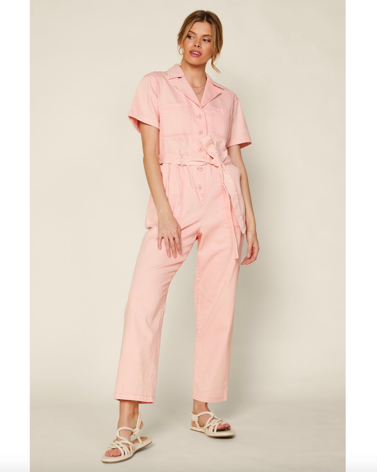 Model wearing skies are blue utility jumpsuit in peach wearing white shoes on a beige background