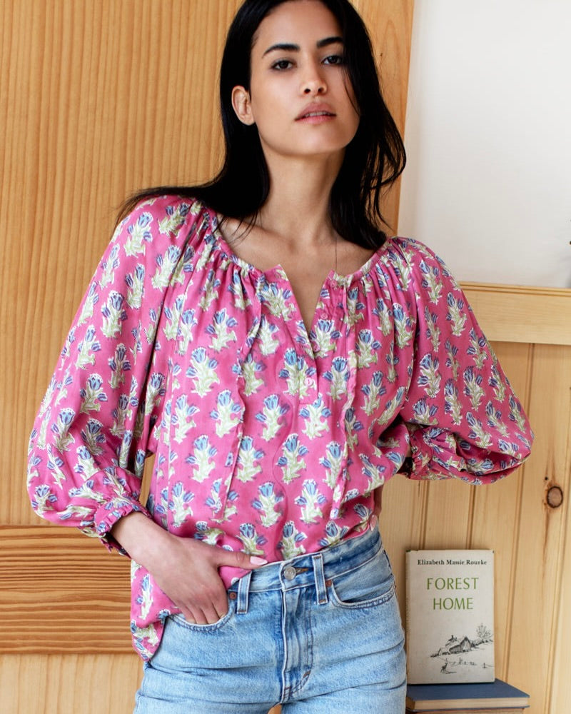 Woman standing wearing blue and green floral with pink background blouse with jeans against a wooden wall.